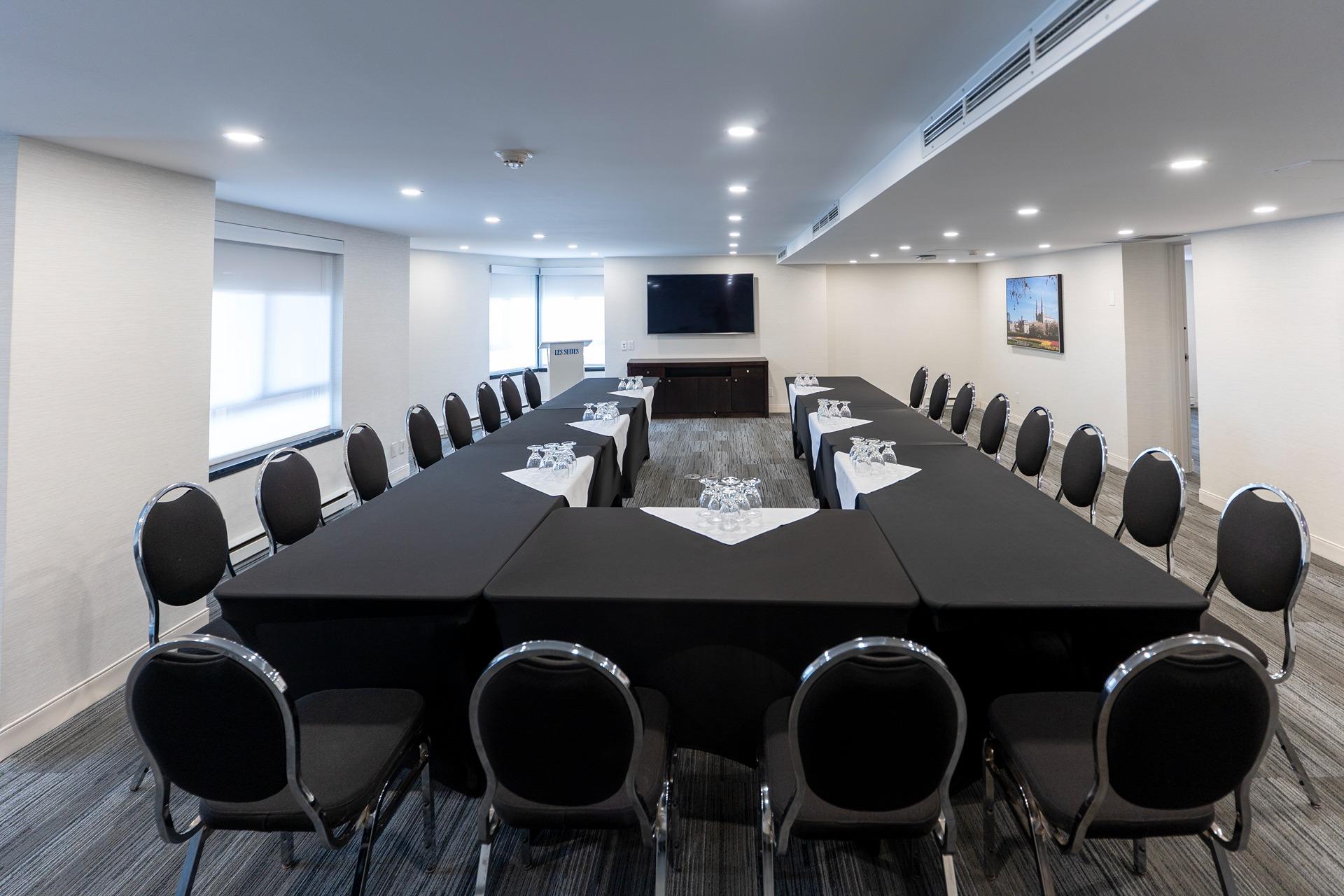 Conference room with U-shaped black-clothed tables, glasses, and chairs facing a wall-mounted TV on the far wall.