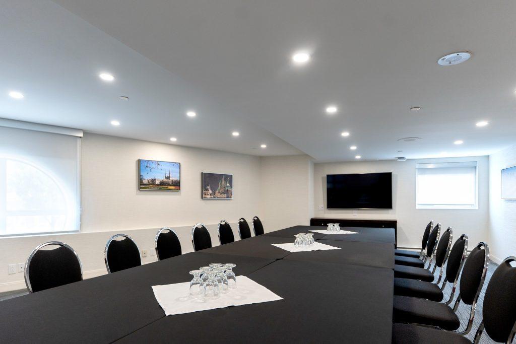 Conference room with a long black-table setup, glasses, and black chairs; wall-mounted TV and framed art on beige walls.